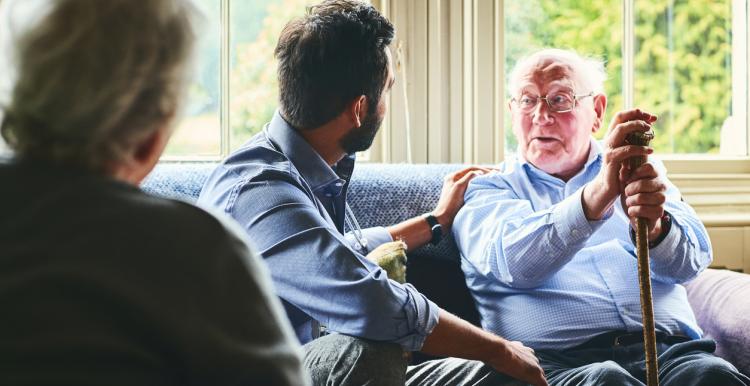 A man leans on a stick and talks to two health professionals. A man places a supportive hand on his shoulder.